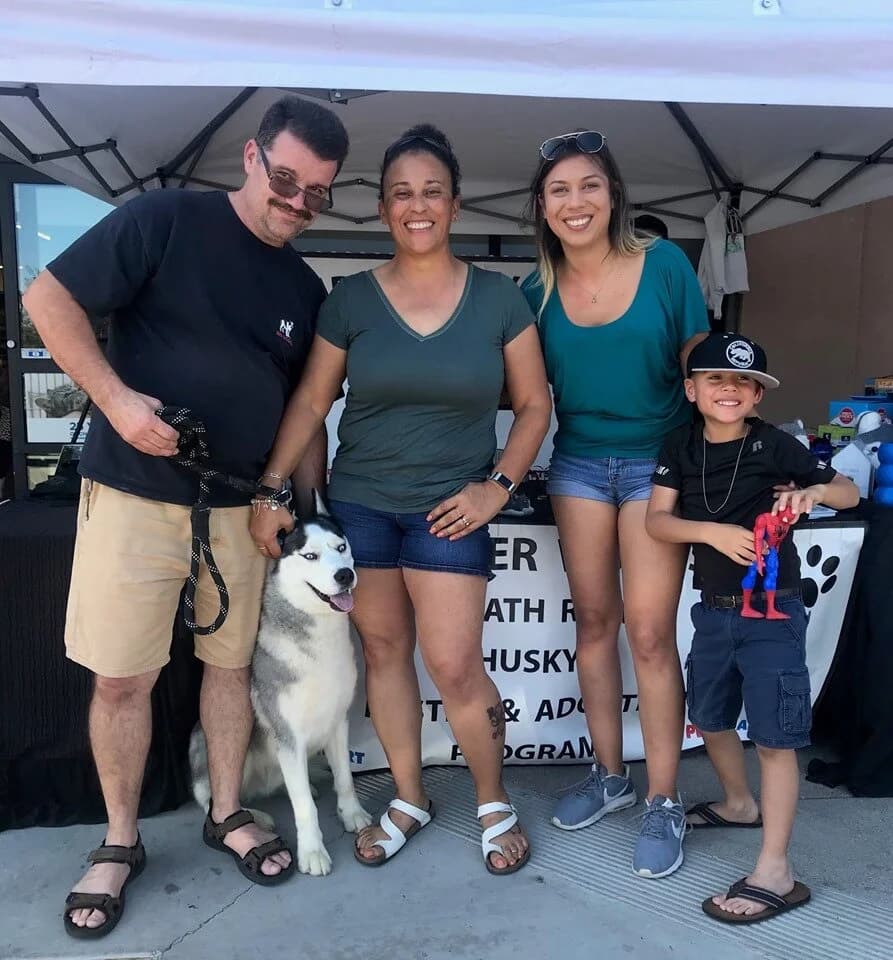 A smiling group poses with a black-and-white husky at a rescue organization adoption event booth