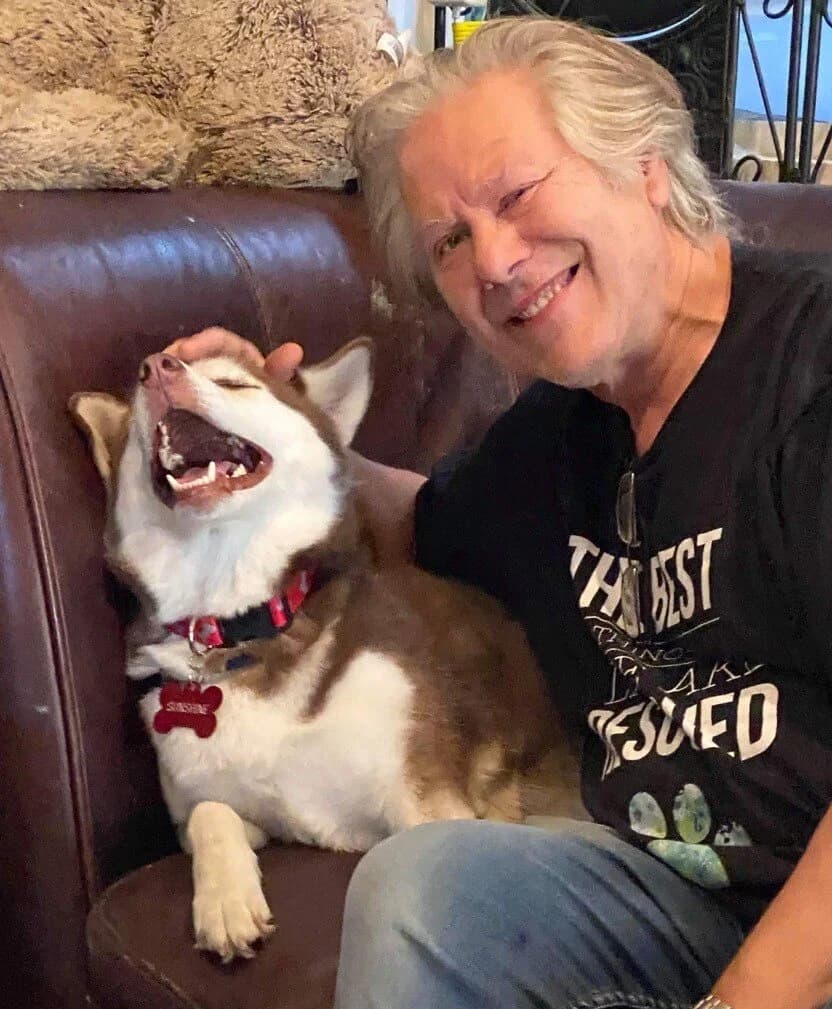 An older man laughing joyfully while a happy red and white Husky named Sunshine yawns beside him