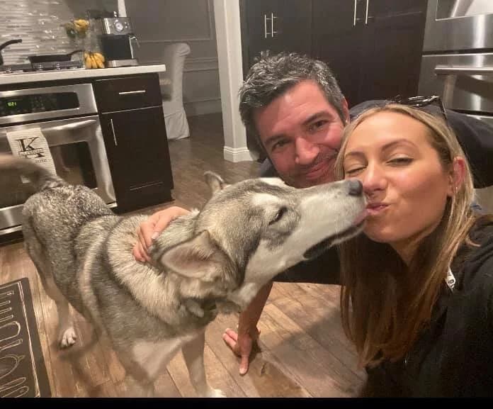 A couple smiling in a kitchen as a grey Husky gives one of them a kiss
